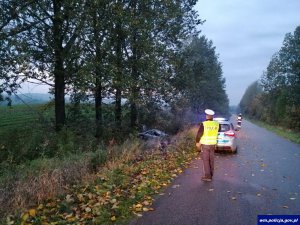 stojący na jezdni policjant, w tle radiowóz, po lewej stronie samochód leżący na dachu